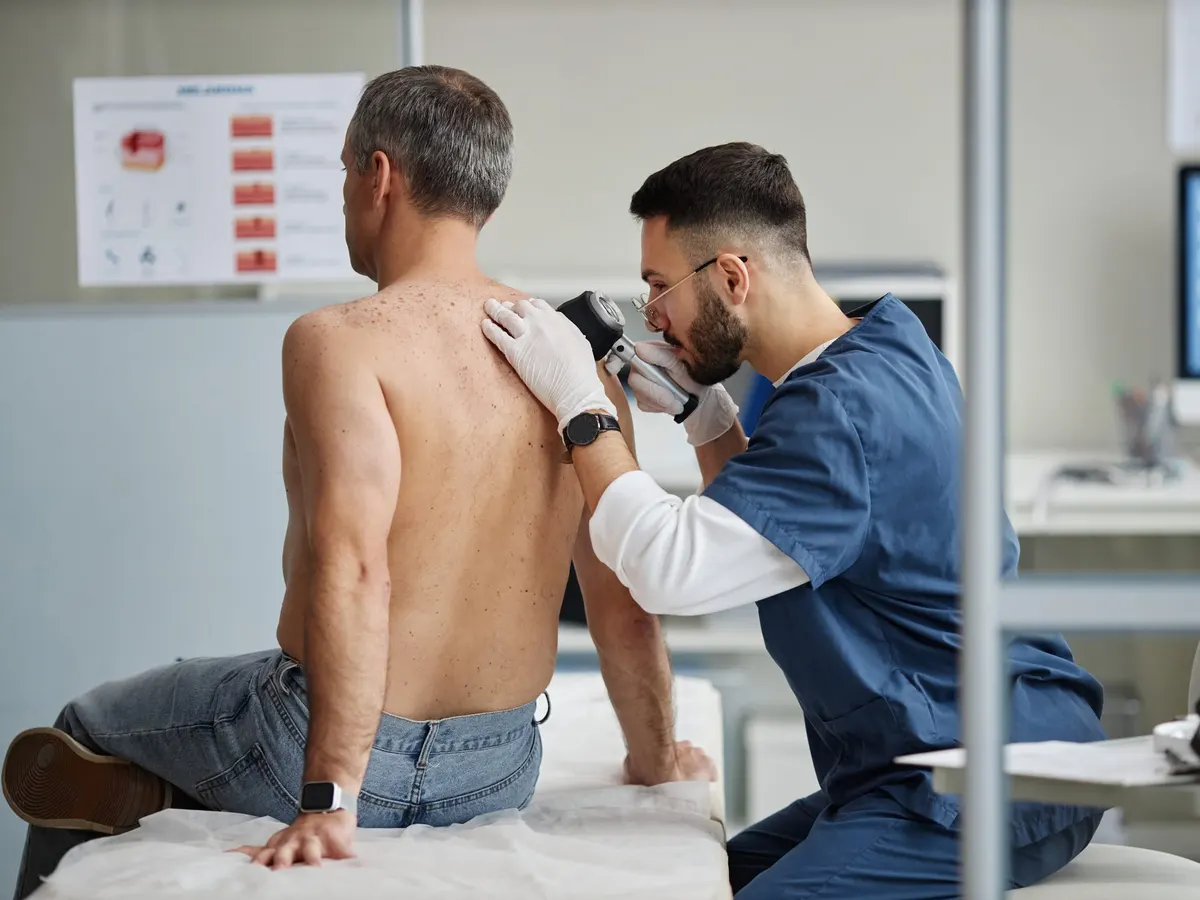 Stock photograph of models: a clinician in blue scrubs examines a seated man's bare back with a dermatoscope