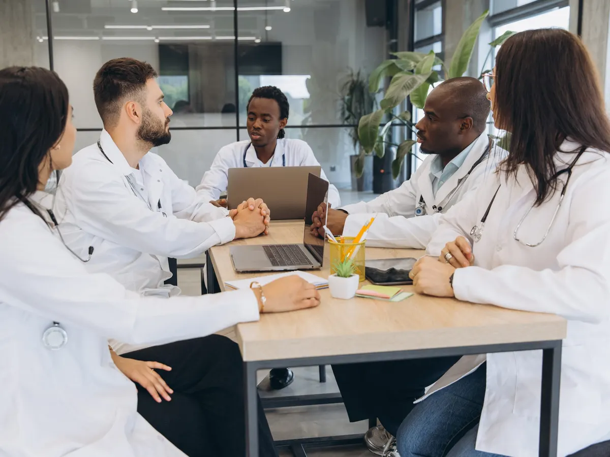 Stock photograph of models: five doctors in white coats talking around a meeting table with laptops