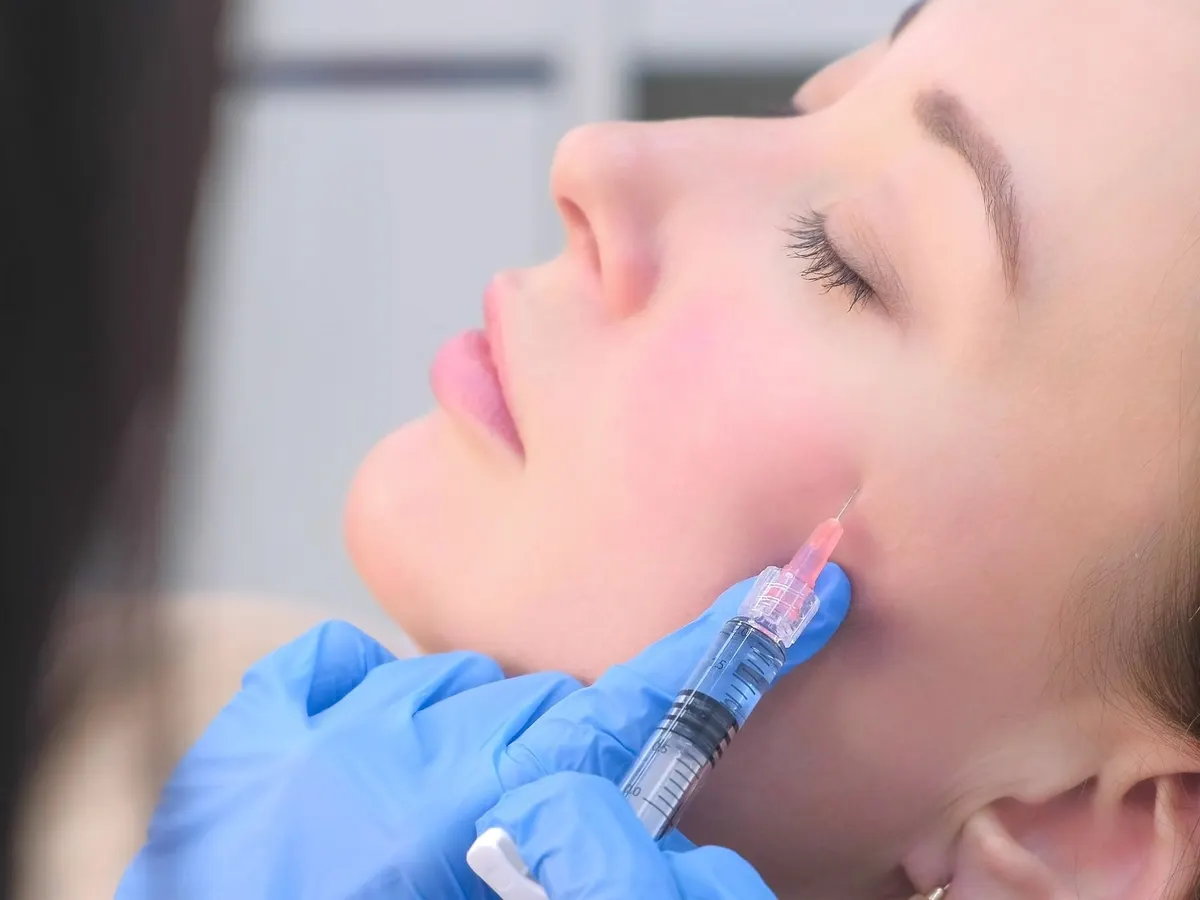Stock photograph of a model: a blue-gloved hand injects the lower cheek of a woman lying back with her eyes closed