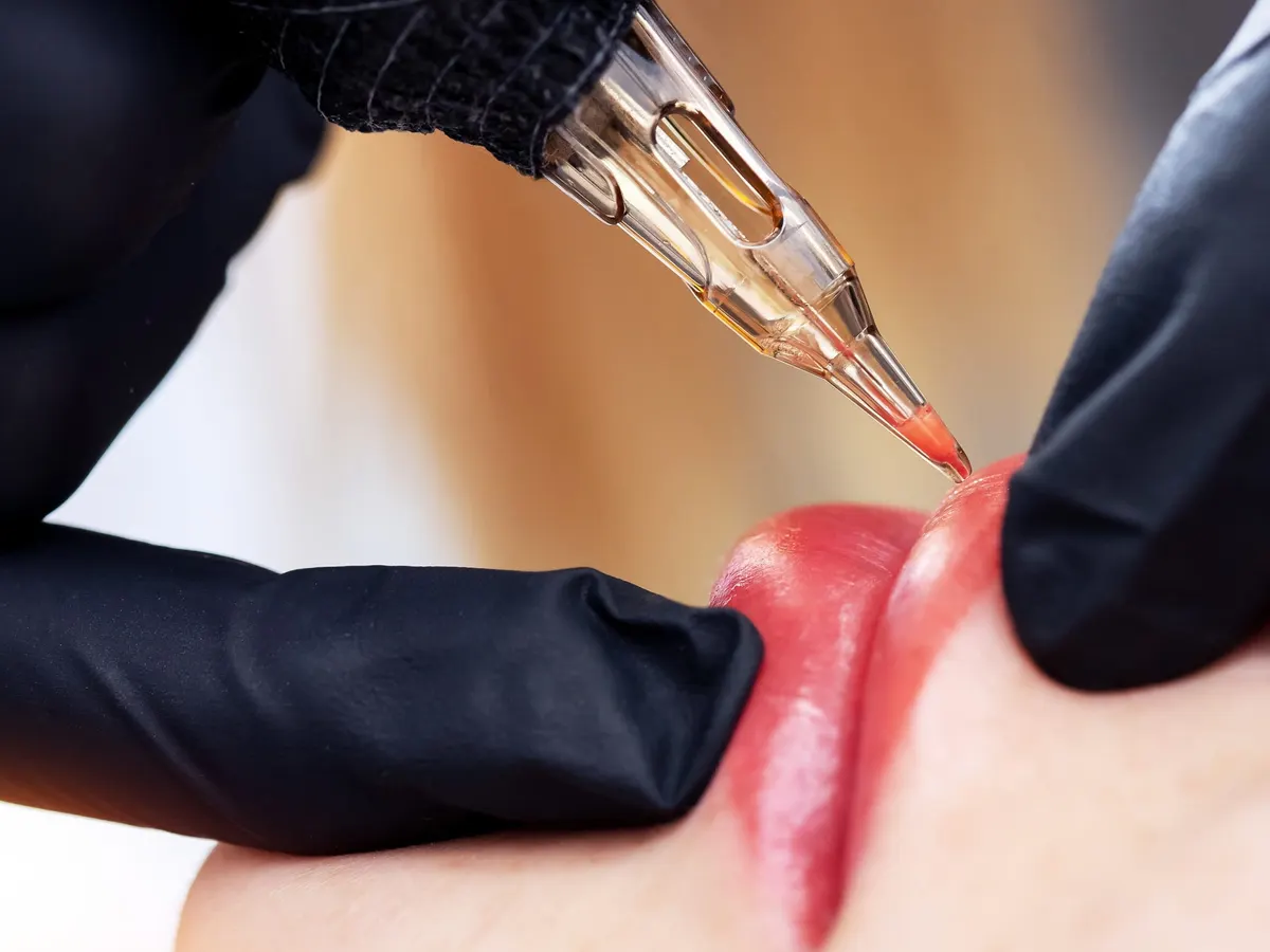Stock photograph of a model: black-gloved hands apply red pigment to a woman's lip line with a fine tattoo needle