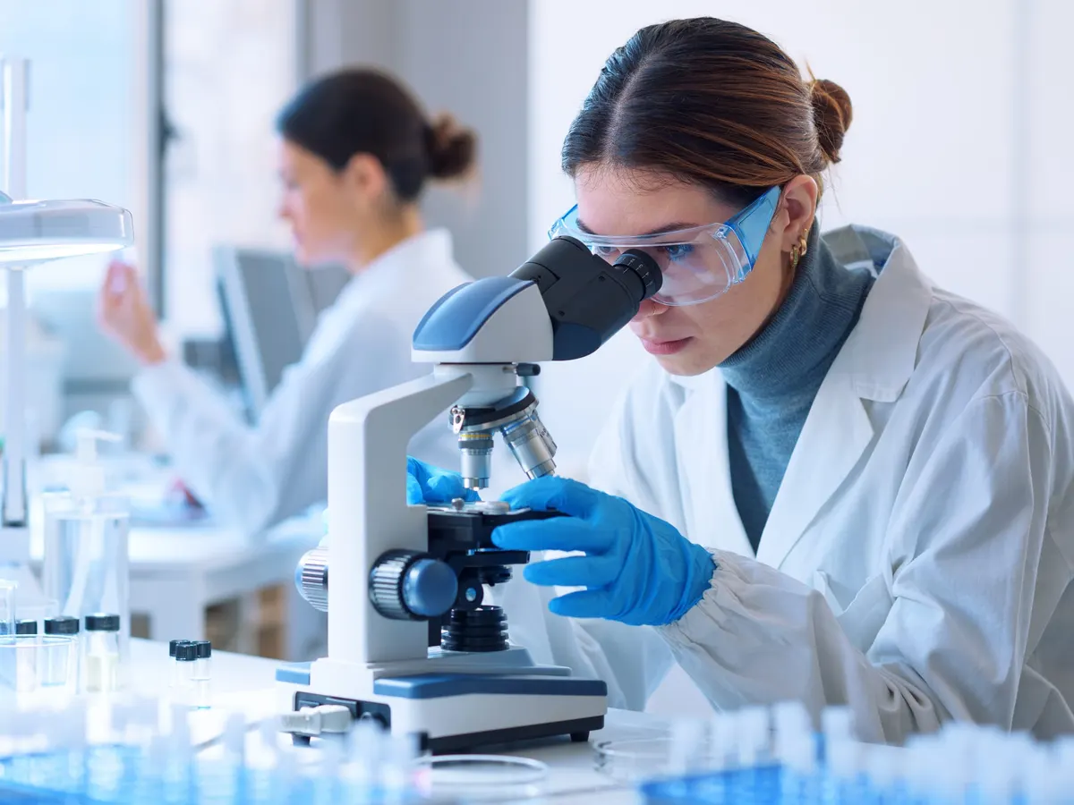 Stock photograph of models: a researcher in safety glasses looks into a microscope in a bright laboratory