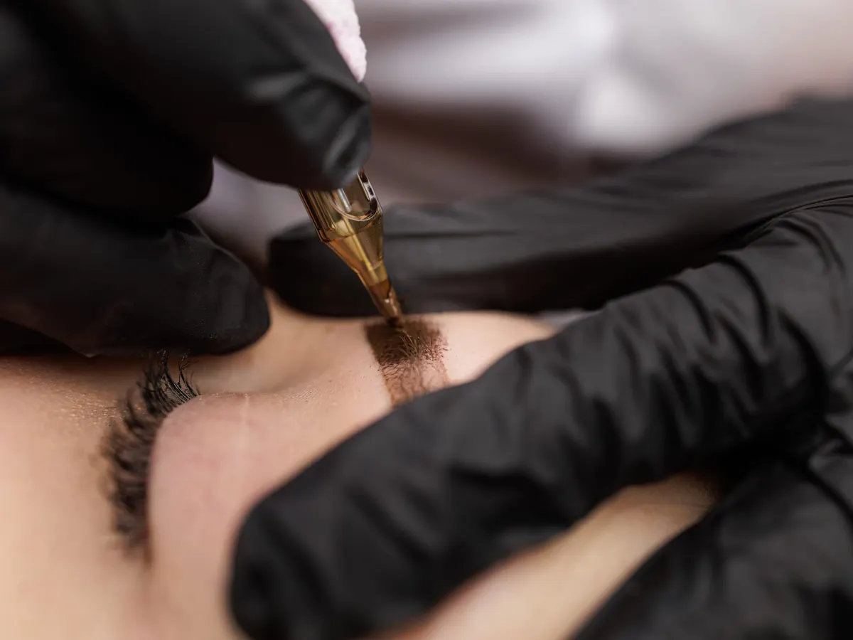 Stock photograph of a model: black-gloved hands guide a fine pigment needle along an eyebrow, close up