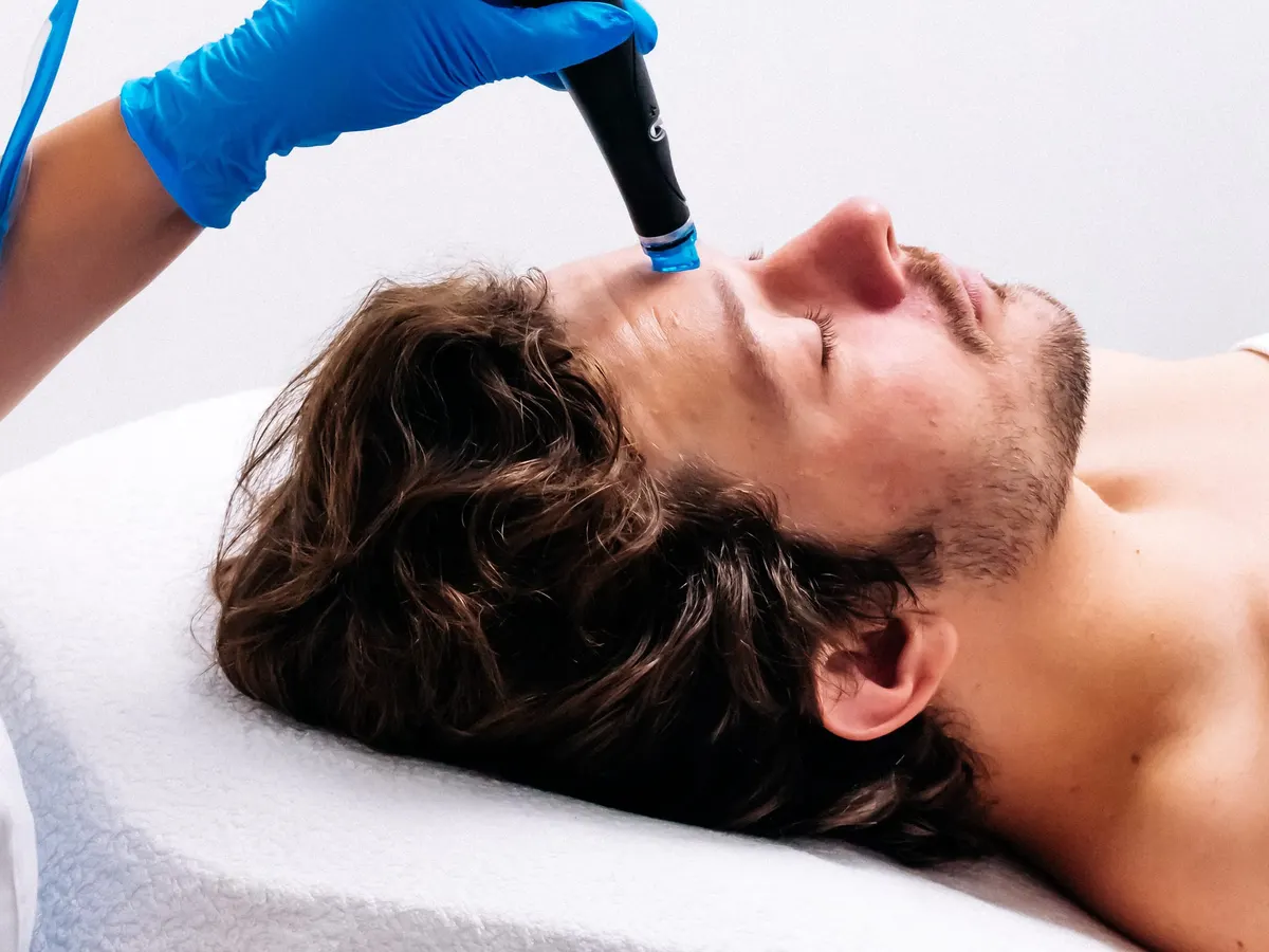 Stock photograph of a model: a man lies back with eyes closed as a gloved esthetician runs a hydrafacial tip over his forehead