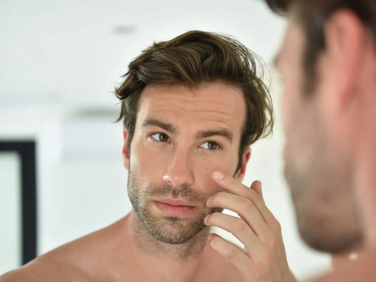 Stock photograph of a model: a man touches the skin under his eye while looking into a bathroom mirror
