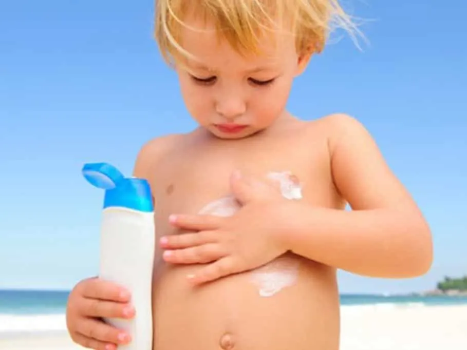 A model toddler on a beach rubbing sunscreen on his tummy, holding a bottle