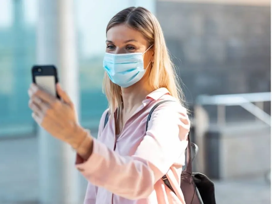 A model wearing a face mask taking a selfie with her phone outdoors