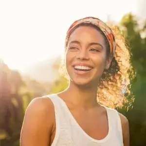 A model smiling outdoors in warm sunlight, wearing a patterned headband and white tank top