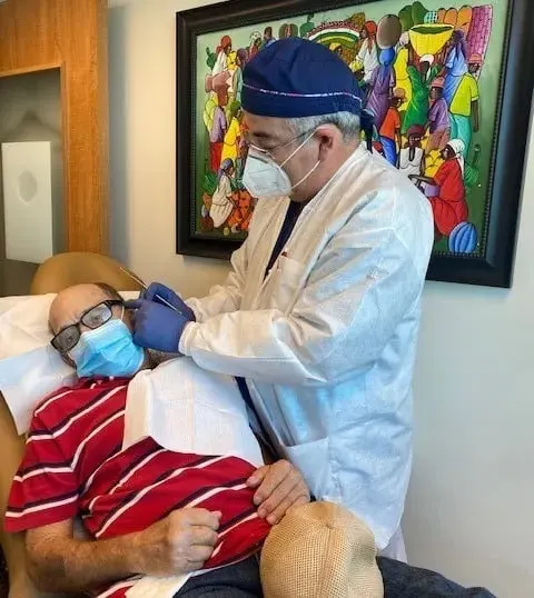 A physician in a surgical cap and mask treating a reclining patient in a striped red shirt