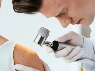 A dermatologist examining a woman's back and shoulder with a handheld dermatoscope
