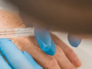 A gloved hand holding a syringe against the back of a hand with freckled skin