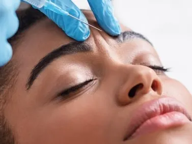 A model with eyes closed receiving an injection near the forehead from a blue-gloved hand