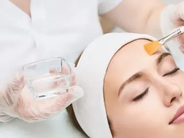 A model in a head wrap lying back while a gloved clinician applies a chemical peel with a brush