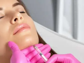 A woman lying back with eyes closed receiving an injection along the jawline from pink-gloved hands