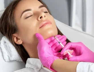 A woman lying back with eyes closed receiving an injection along the jawline from pink-gloved hands
