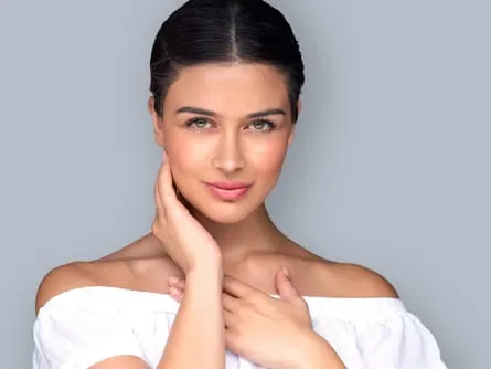 A smiling model with dark hair pulled back, one hand at her neck, in a white off-shoulder top