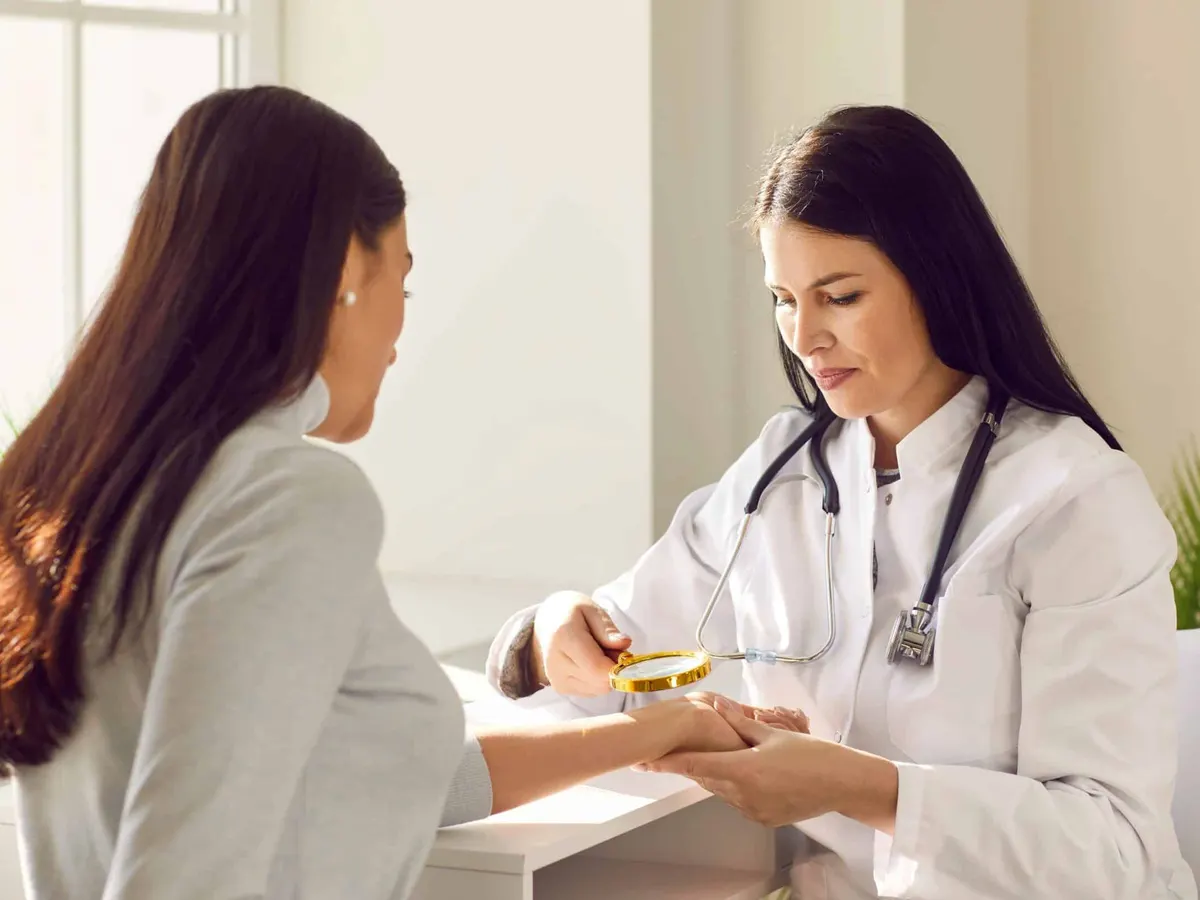 A dermatologist in a white coat examining a model's hand with a magnifying glass
