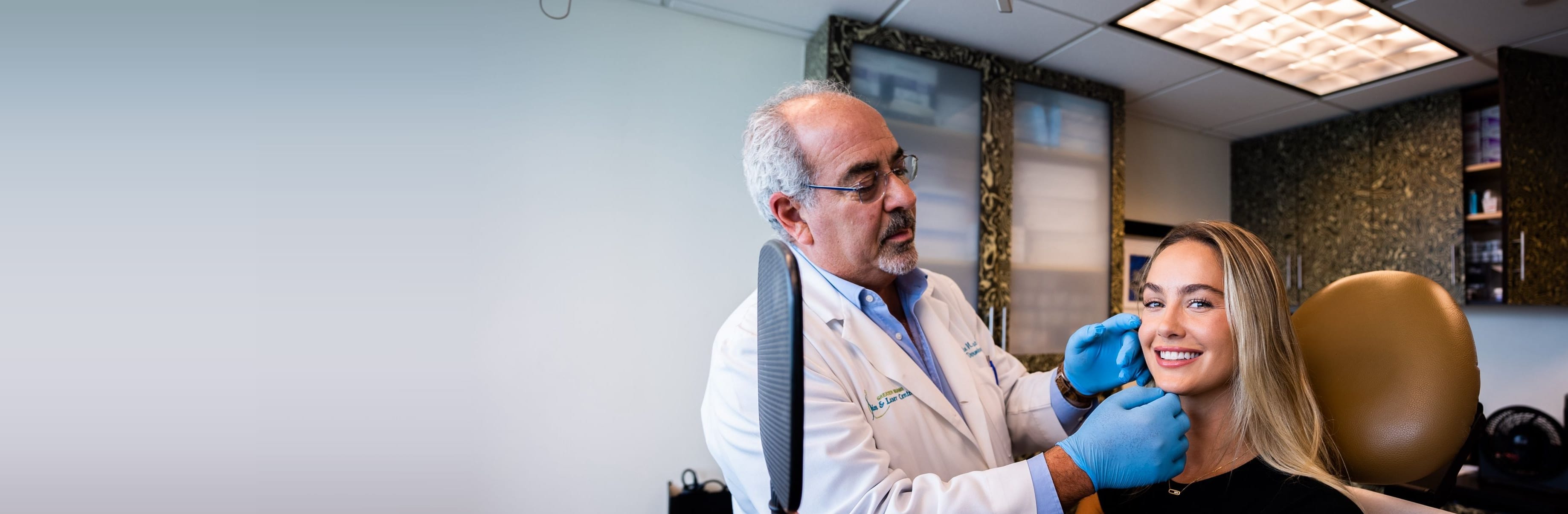 Dr. Martin N. Zaiac, in a white coat and blue gloves, examines a smiling woman's jawline as she holds a hand mirror in a treatment room at Greater Miami Skin & Laser Center