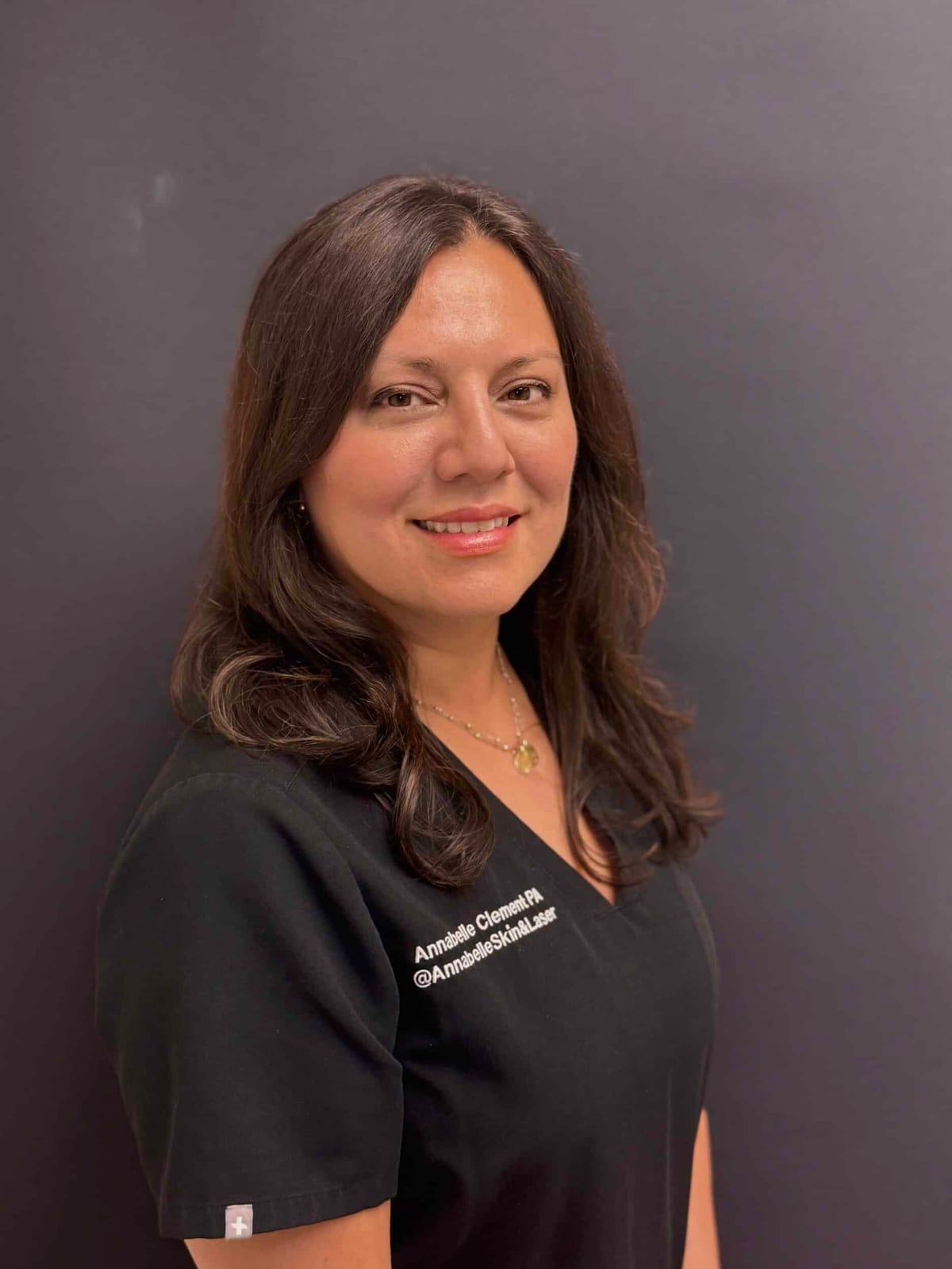Annabelle Clement, PA-C, smiling, with long dark hair, in black scrubs against a grey backdrop
