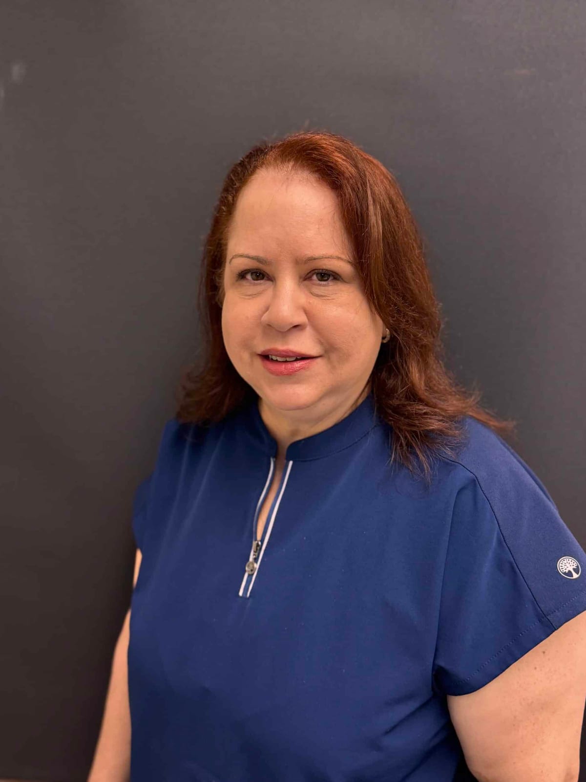 Nellie Cortez, aesthetician, smiling, with auburn hair, in a navy top against a grey backdrop