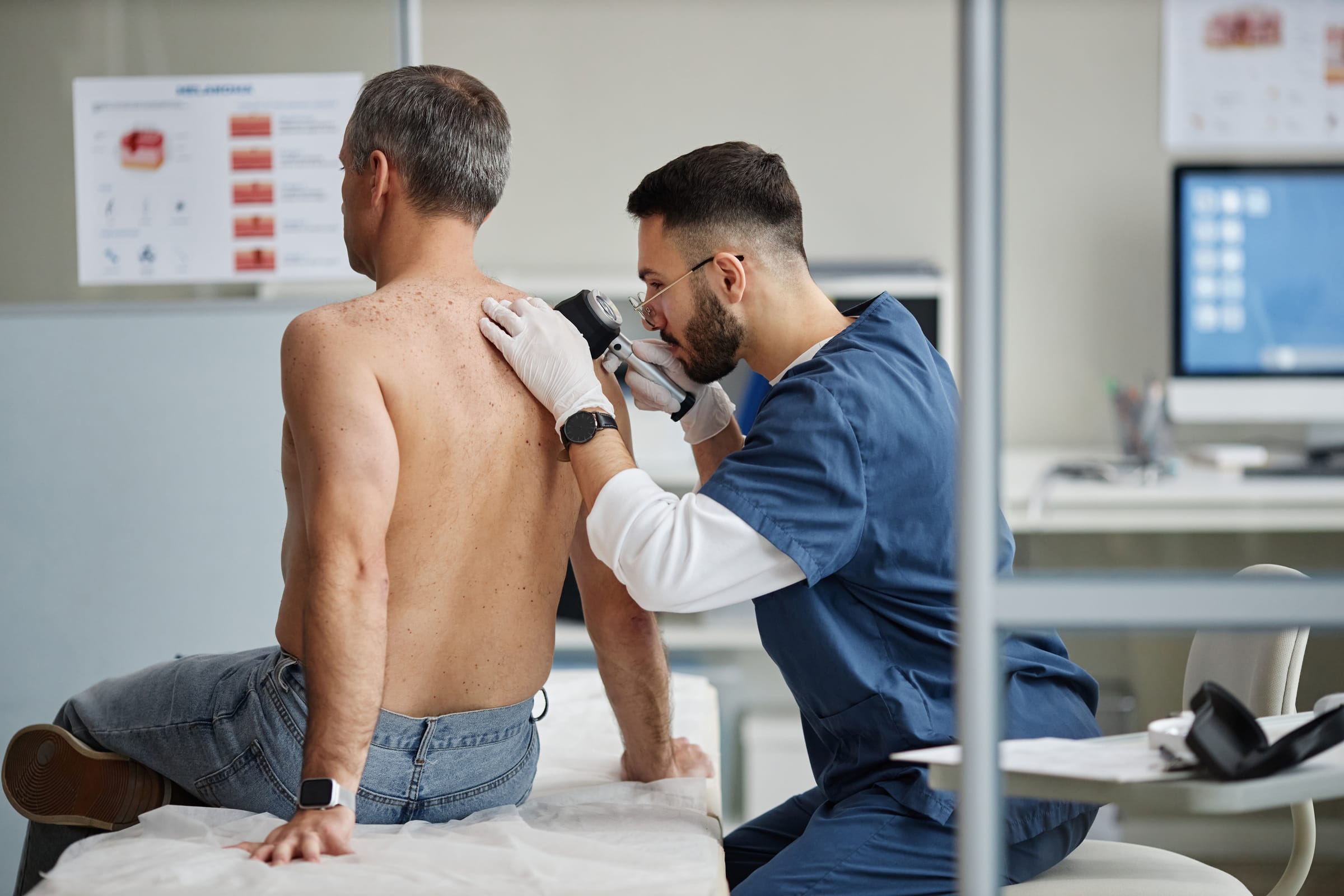 Stock photograph of models: a clinician in blue scrubs examines a seated man's bare back with a dermatoscope