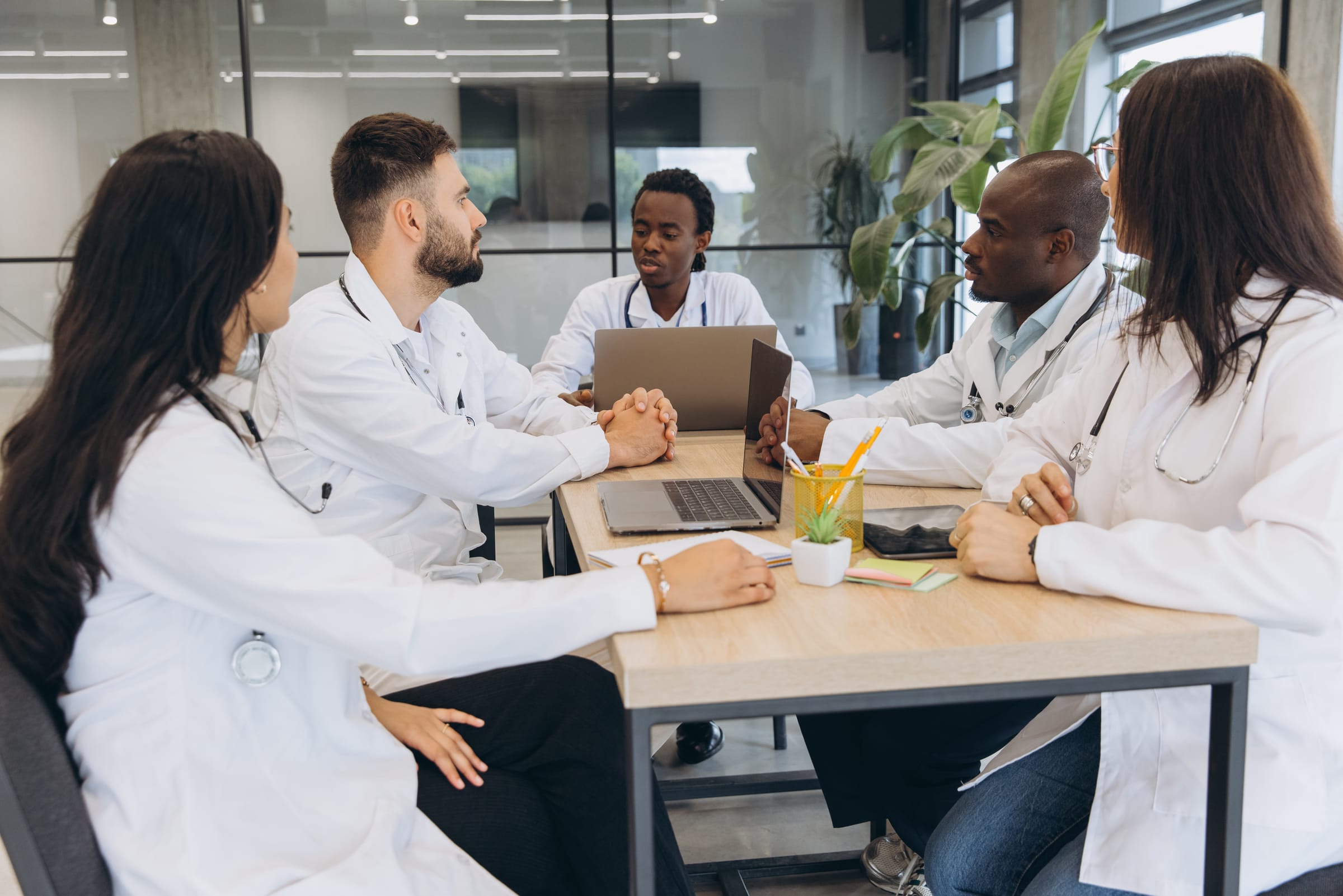 Stock photograph of models: five doctors in white coats talking around a meeting table with laptops