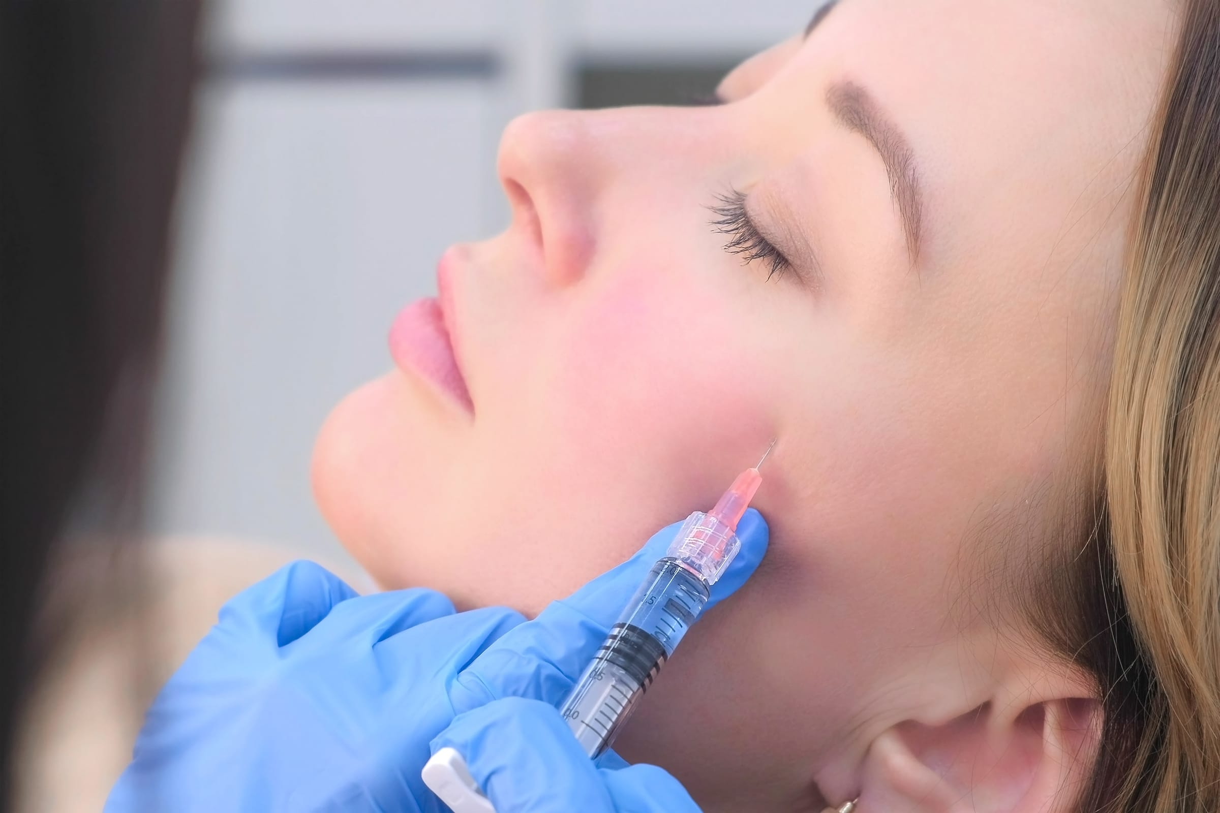 Stock photograph of a model: a blue-gloved hand injects the lower cheek of a woman lying back with her eyes closed