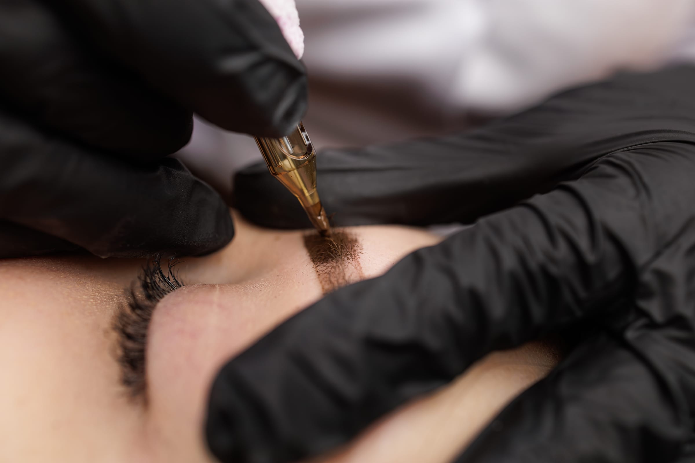 Stock photograph of a model: black-gloved hands guide a fine pigment needle along an eyebrow, close up