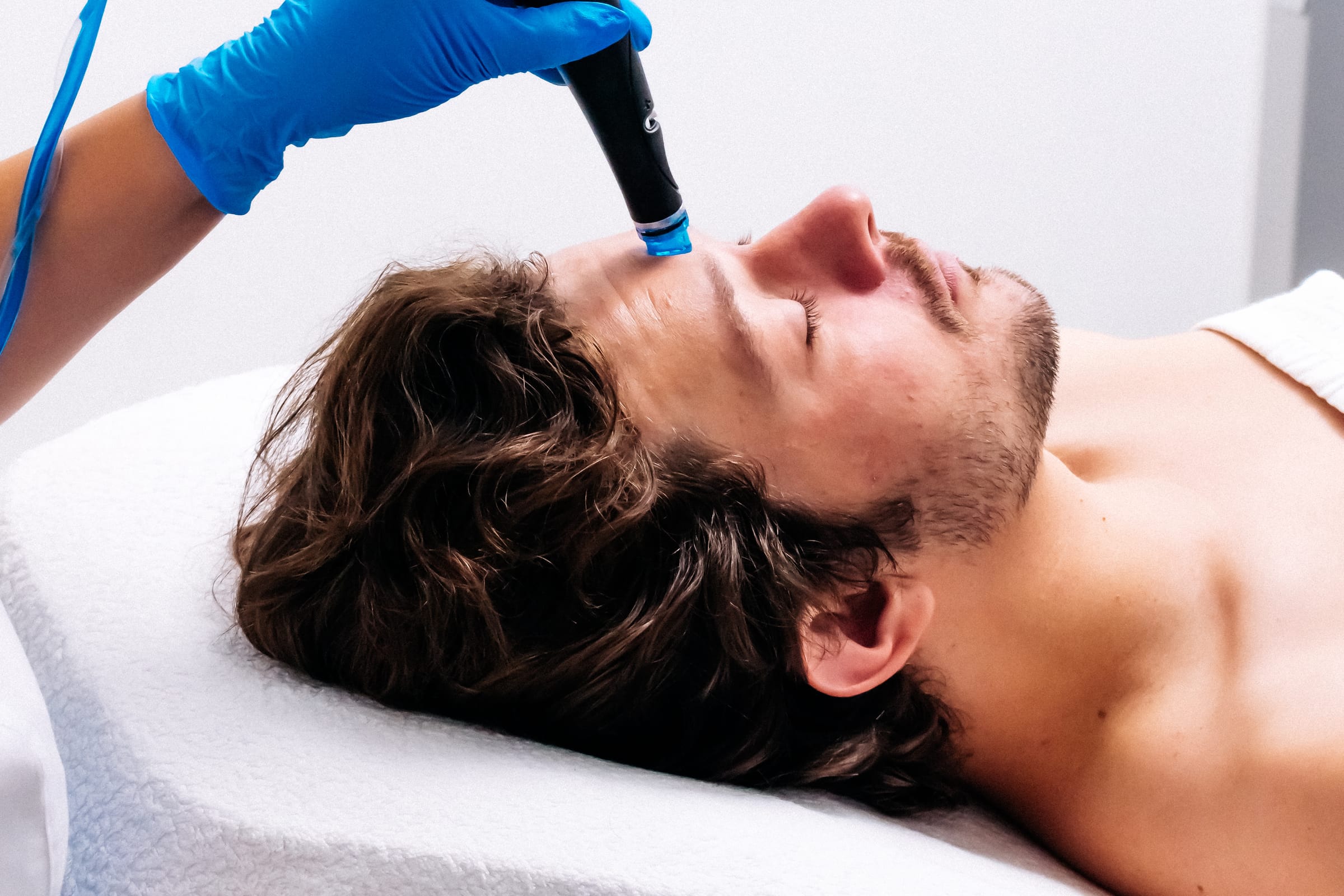 Stock photograph of a model: a man lies back with eyes closed as a gloved esthetician runs a hydrafacial tip over his forehead