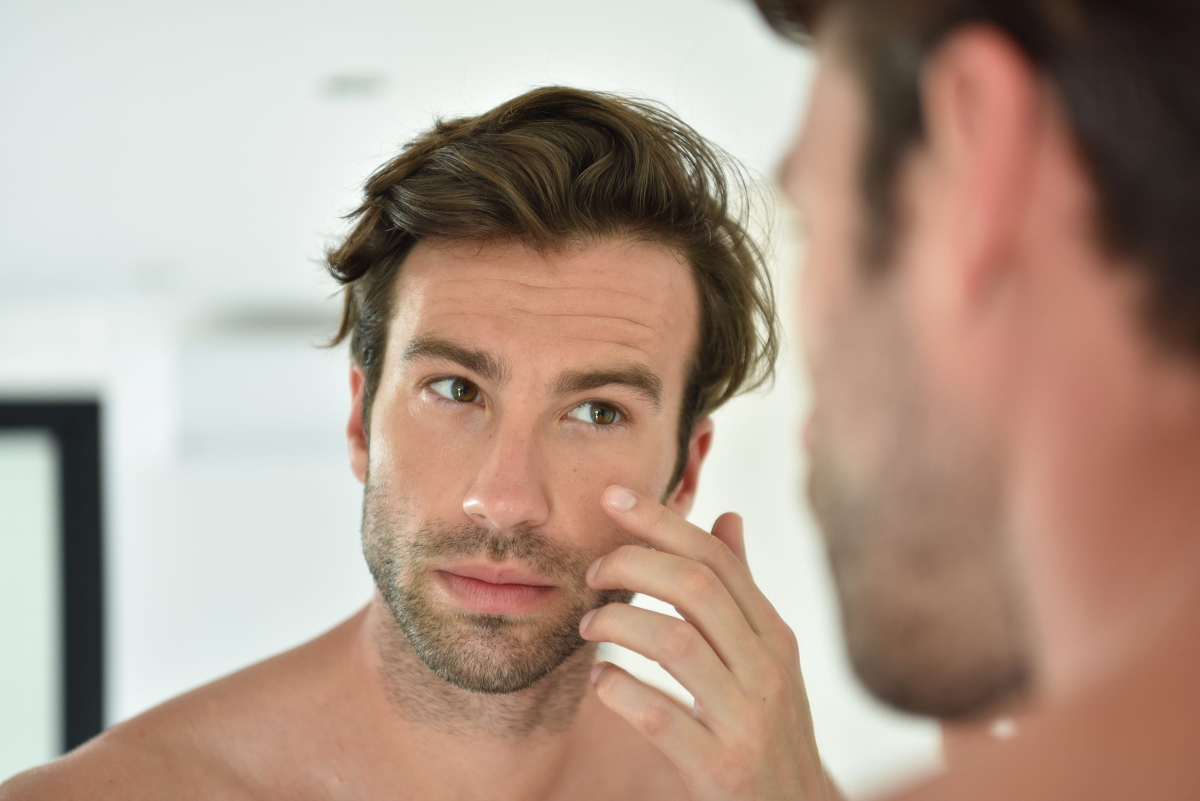Stock photograph of a model: a man touches the skin under his eye while looking into a bathroom mirror
