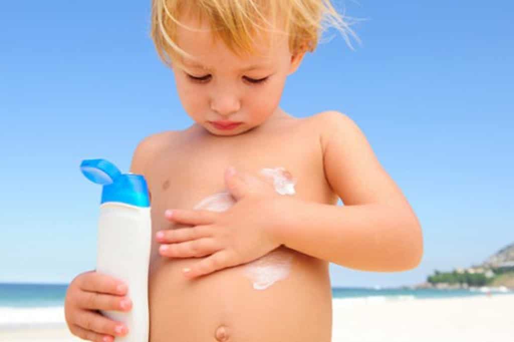 A model toddler on a beach rubbing sunscreen on his tummy, holding a bottle
