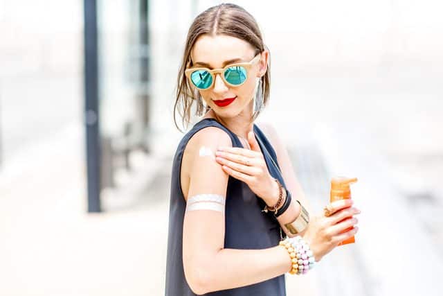 A model in sunglasses applying sunscreen to her shoulder, holding an orange bottle