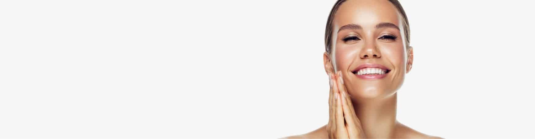 A smiling model with her hair pulled back touching her cheek, on a pale grey background