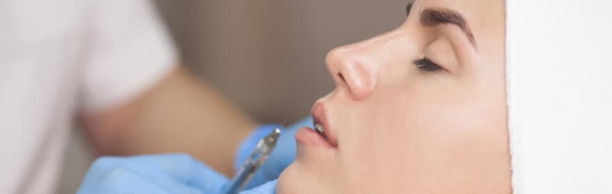 A model with her eyes closed receiving a lip filler injection from a gloved hand