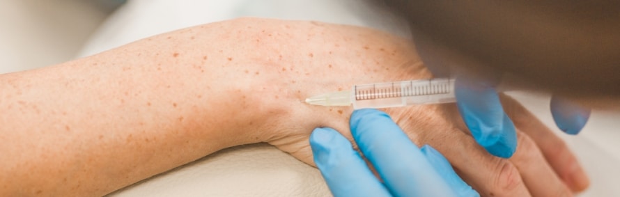 A gloved hand holding a syringe against the back of a hand with freckled skin