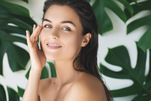 A model with smooth skin touching her face, in front of green tropical leaves