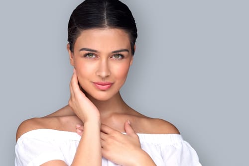 A smiling model with dark hair pulled back, one hand at her neck, in a white off-shoulder top