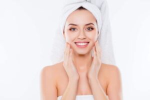 Portrait of stunning woman in towel and turban on head enjoying touching her perfect face skin with fingers isolated on white background | Fraxel Laser Treatment